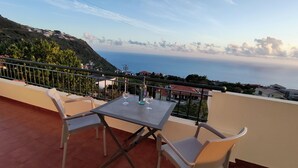 Varanda com mesa e cadeiras, com vista para a costa, casas e uma paisagem montanhosa.
