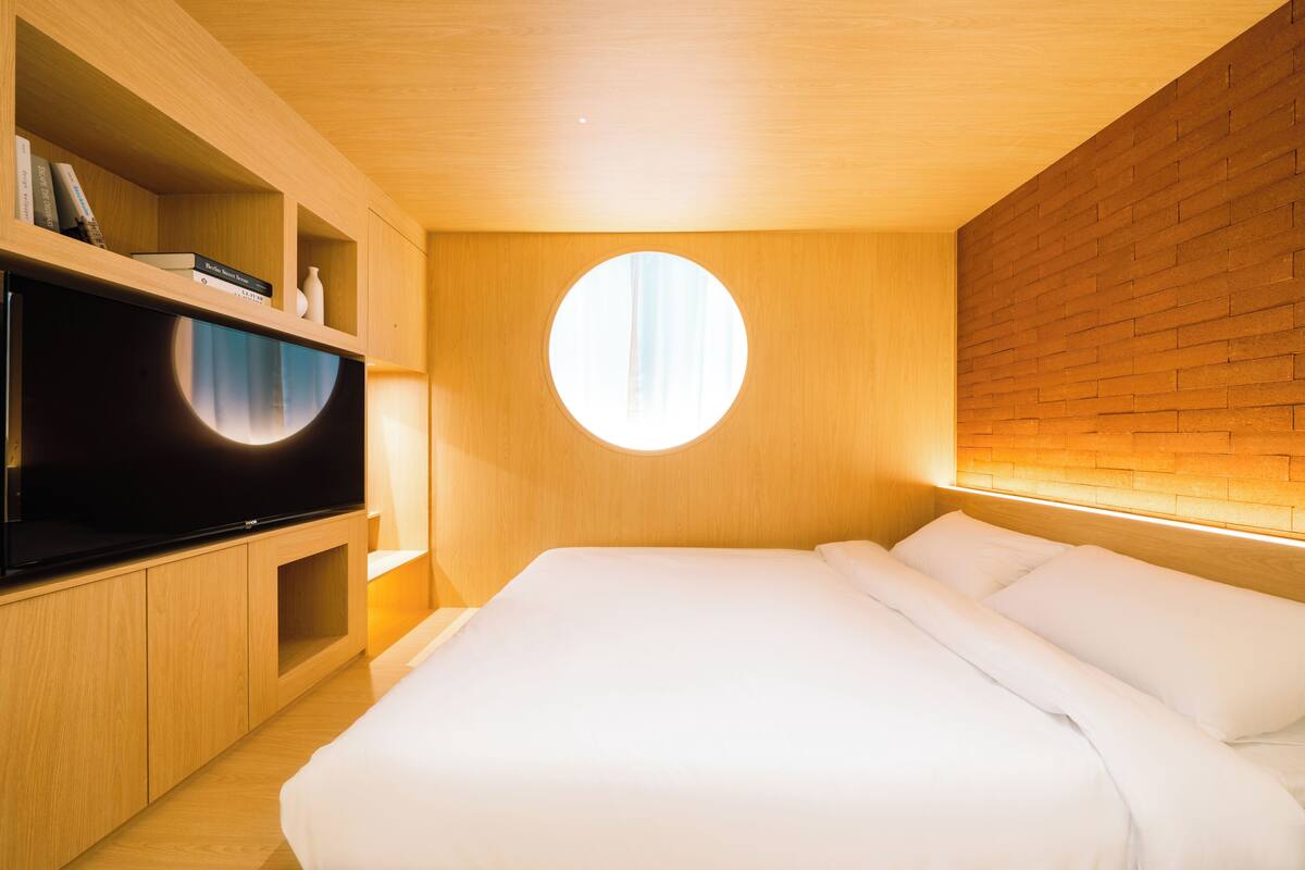 Ein modernes Schlafzimmer mit einem großen Bett, integriertem Fernseher, Holzregalen und einem Rundfenster.