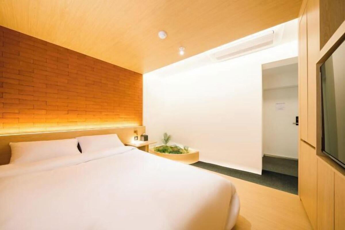 Een moderne hotelkamer met een houten hoofdbord, een groot bed, een nachtkastje met een plant en een aan de muur bevestigde televisie.