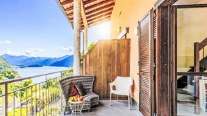 Un balcon agrémenté de meubles en osier, une petite table avec des fruits, et une vue sur les montagnes et l’eau.