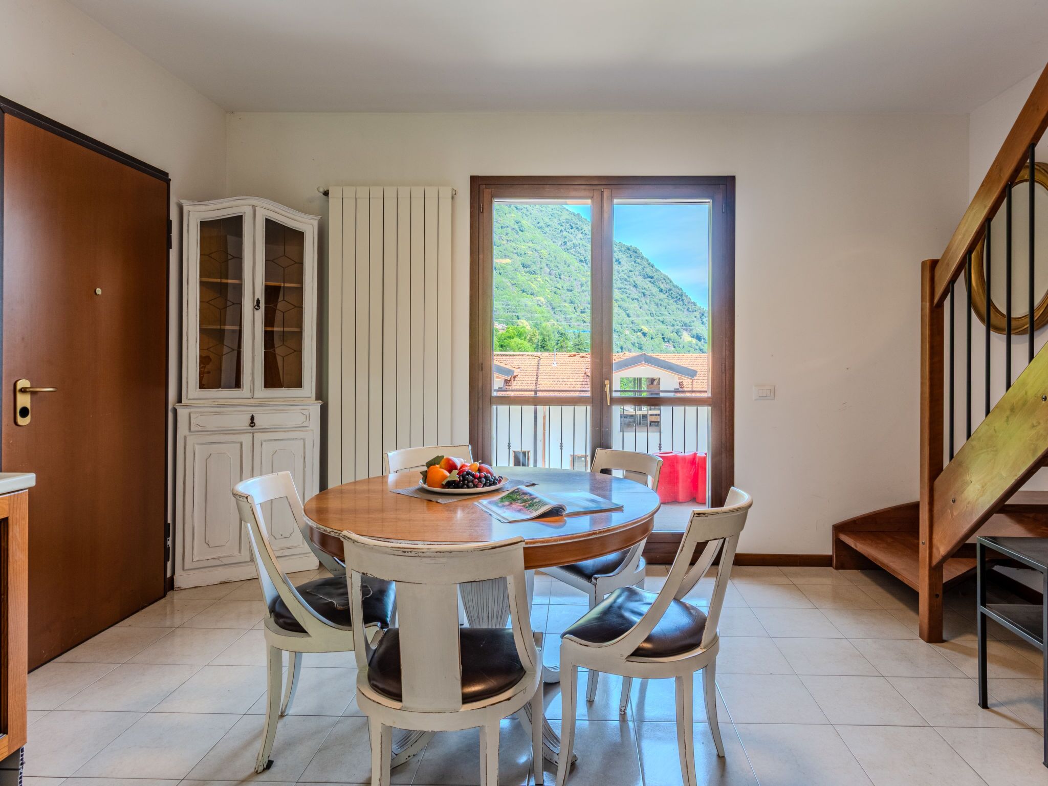 A dining area with a round table, four chairs, a fruit bowl, and a view of a mountainous landscape through a large window.