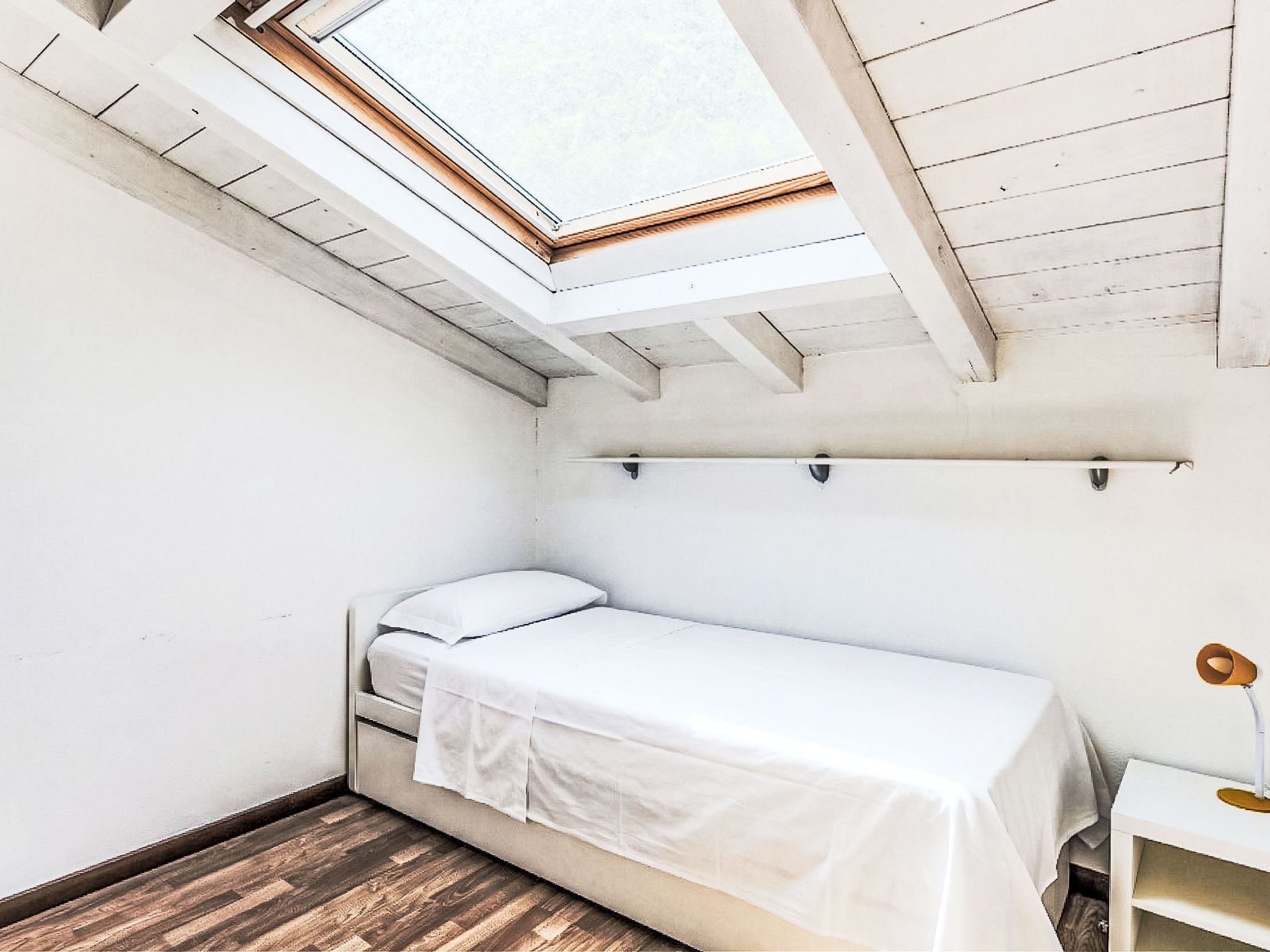 Ein kleines Zimmer mit einem Einzelbett, einem Oberlicht, Holzboden und einem weißen Schreibtisch mit Lampe.