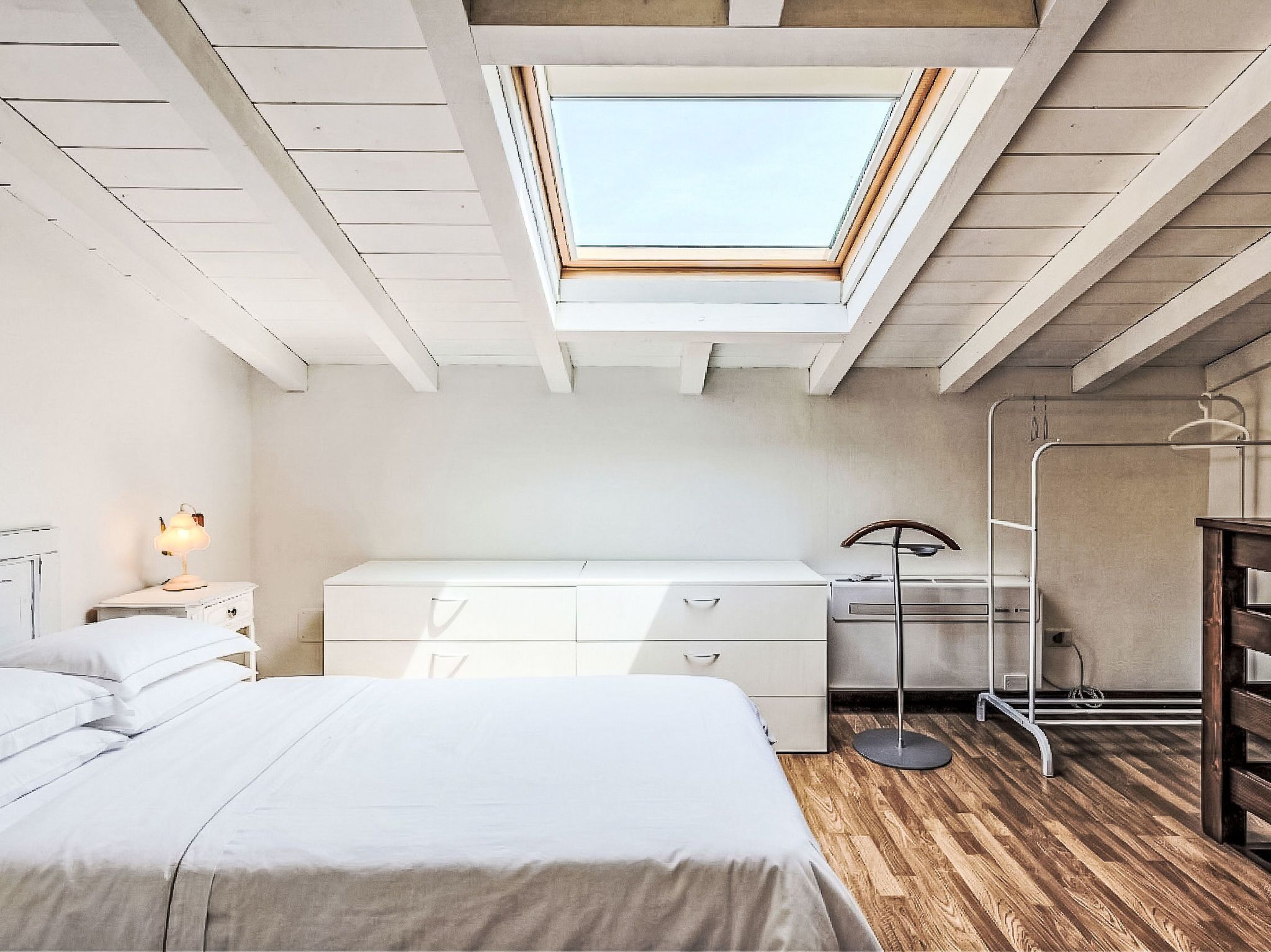 Un dormitorio con tragaluz, una cama, una mesita de noche con lámpara, un aparador y una lámpara de pie.