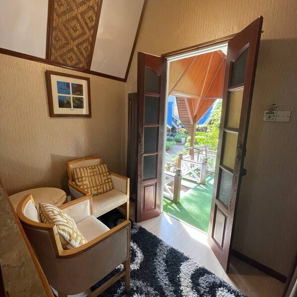 Una habitación acogedora con un sofá de madera, una alfombra blanco y negro y un ventanal con vistas a un jardín y un edificio azul al exterior.