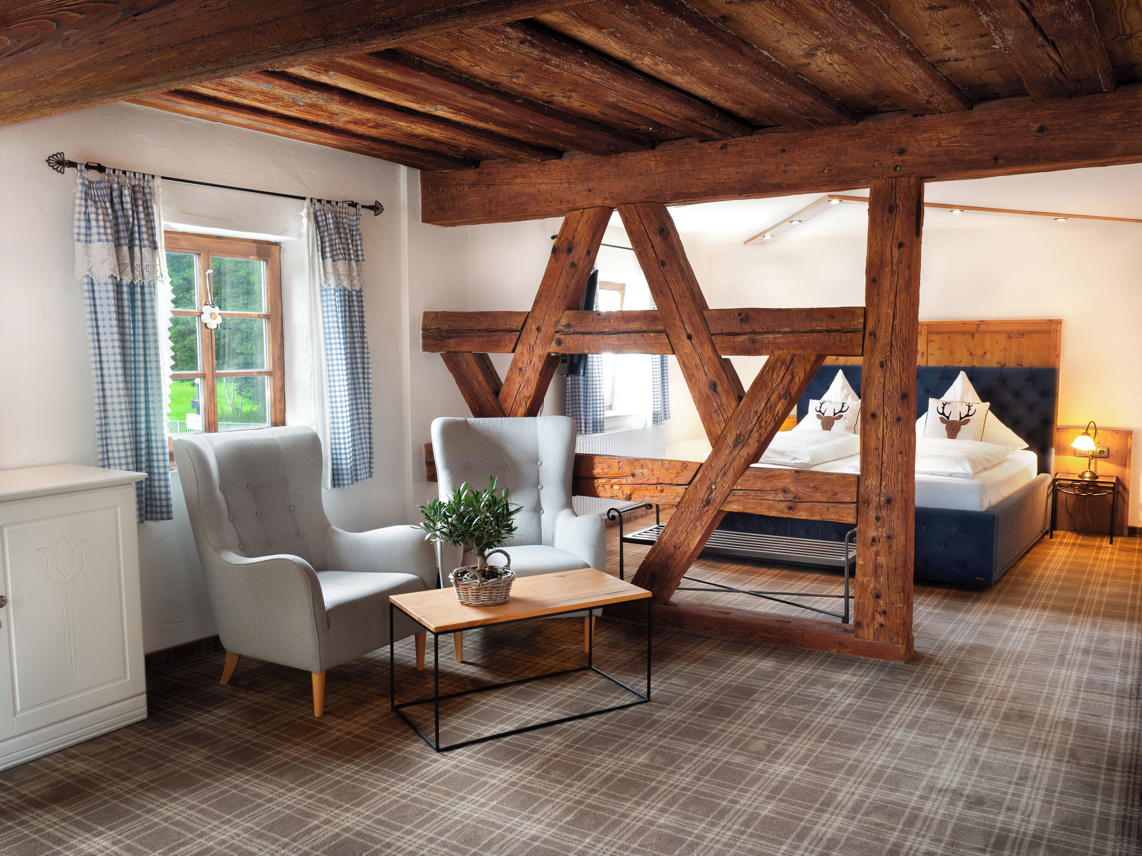 A cozy room with a wooden ceiling, a bed with a blue headboard, a grey armchair, a small table with a plant, and plaid-patterned flooring.