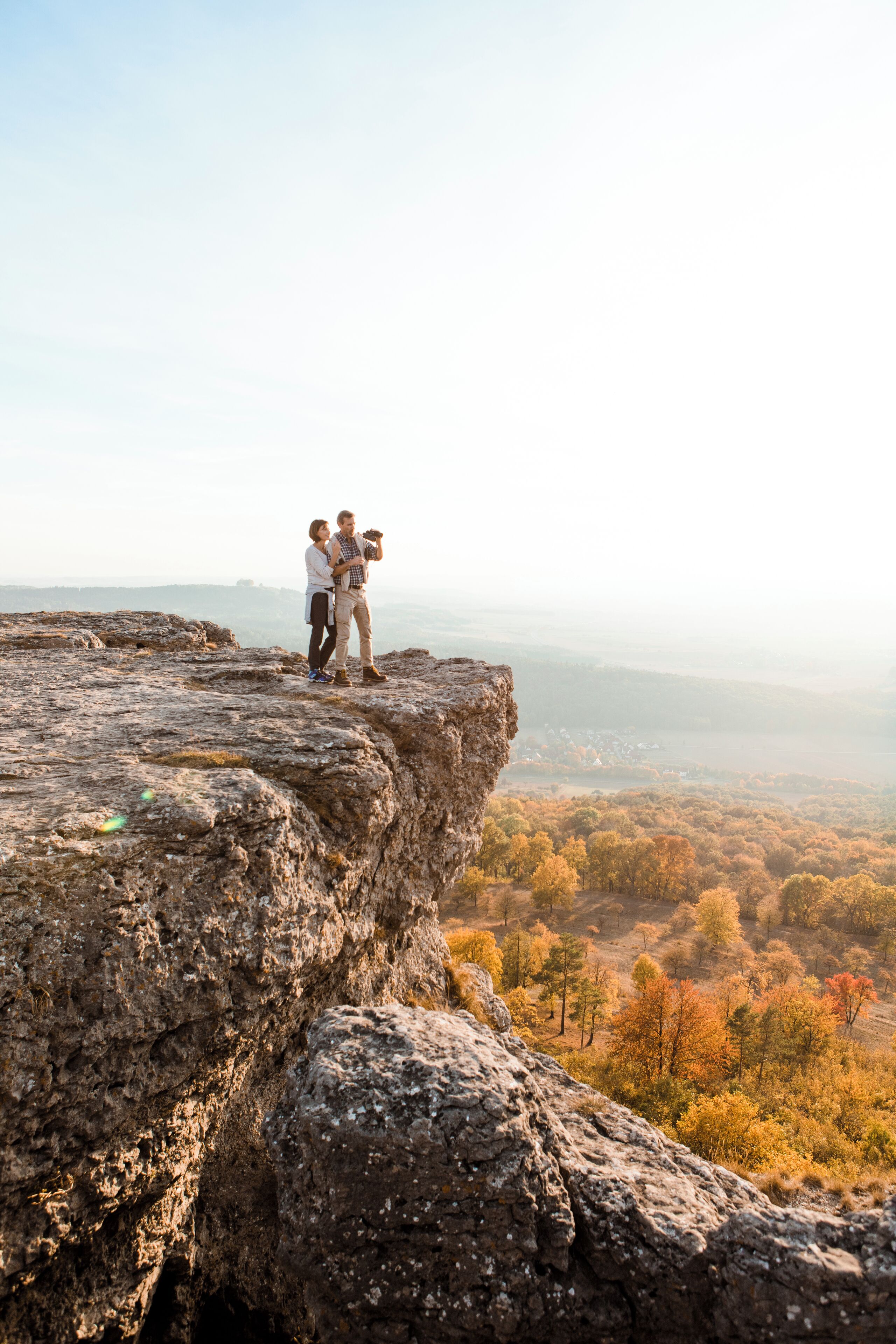 A couple standing on a rocky cliff overlooking a forested valley.