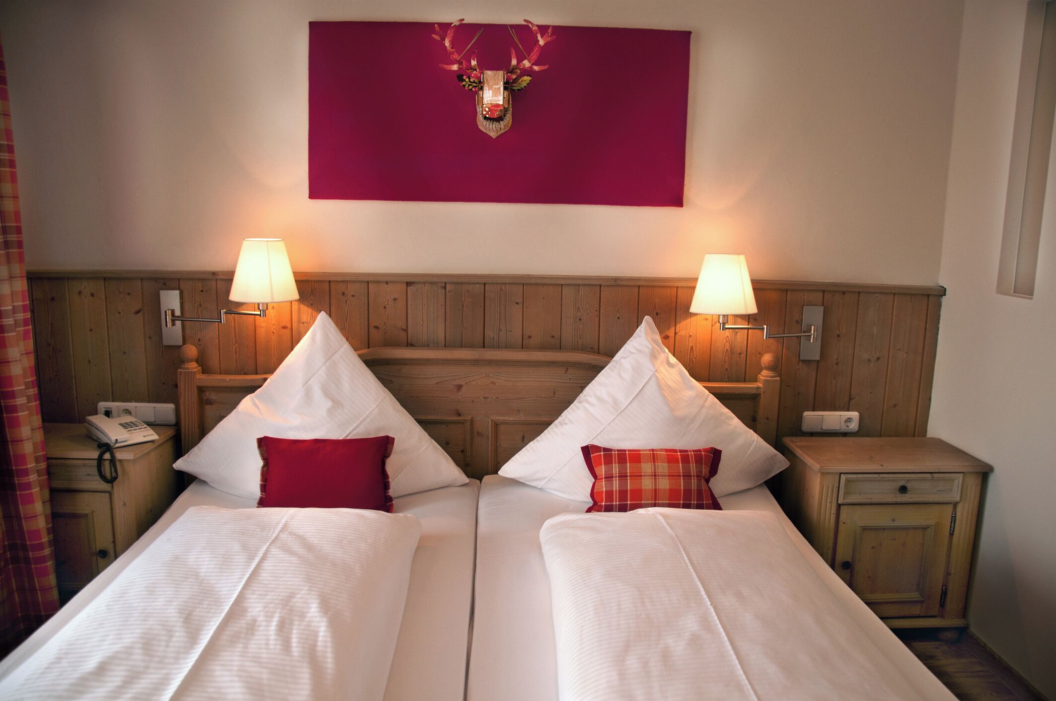 A double bed with white linens and red pillows, two bedside lamps, a wooden headboard, a red wall decoration, and a small bedside table with a phone.