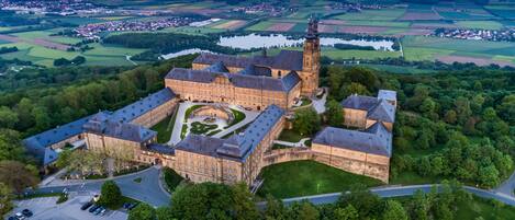 Une vue aérienne d’un ensemble de bâtiments historiques comprenant plusieurs structures, dont une tour imposante, le tout entouré d’une végétation luxuriante et bordé par une rivière.