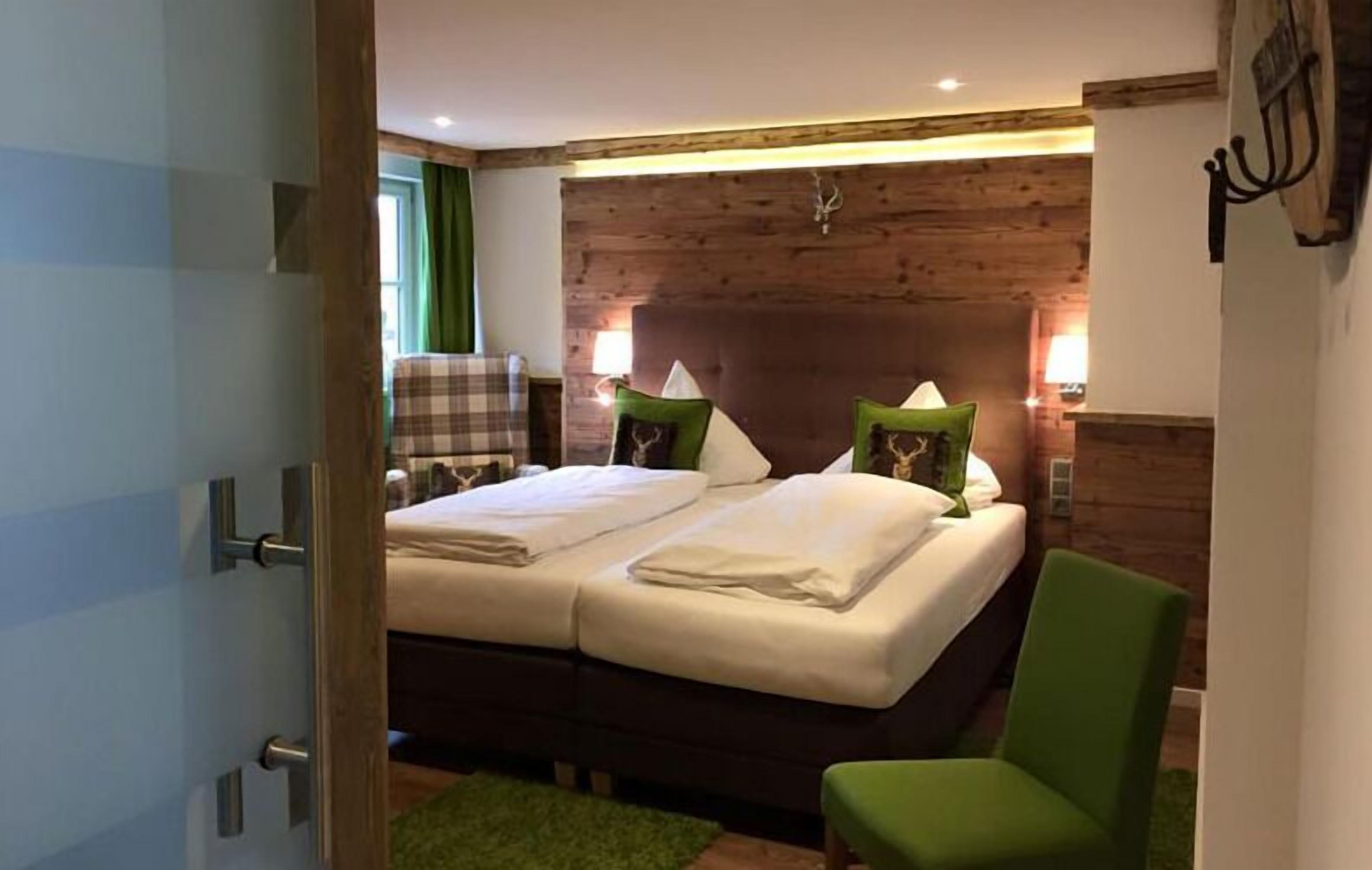 Un dormitorio con cabecera de madera, dos camas con sábanas blancas, una silla verde y una silla a cuadros.