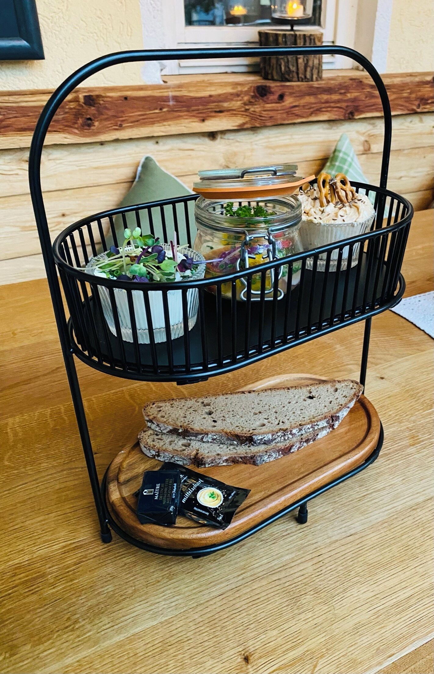 A black metal tiered serving cart with a wooden base, containing a jar of pickles, a jar of mustard, a jar of pickled vegetables, a bowl of salad, and a bowl of pretzels.