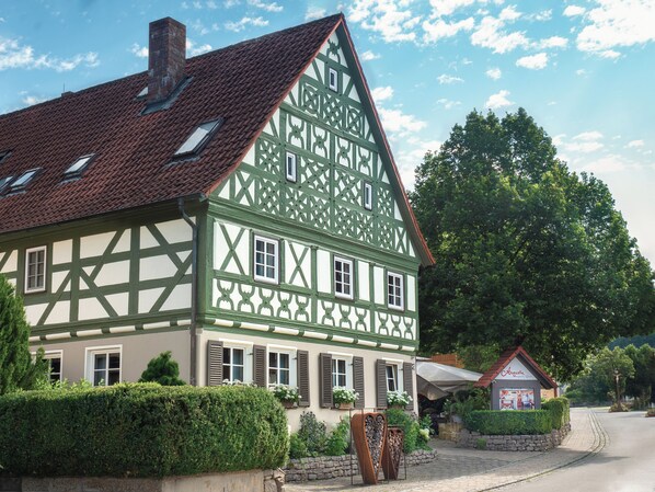 A traditional half-timbered house with a sloped roof, multiple windows, and a small adjacent structure with a sign.
