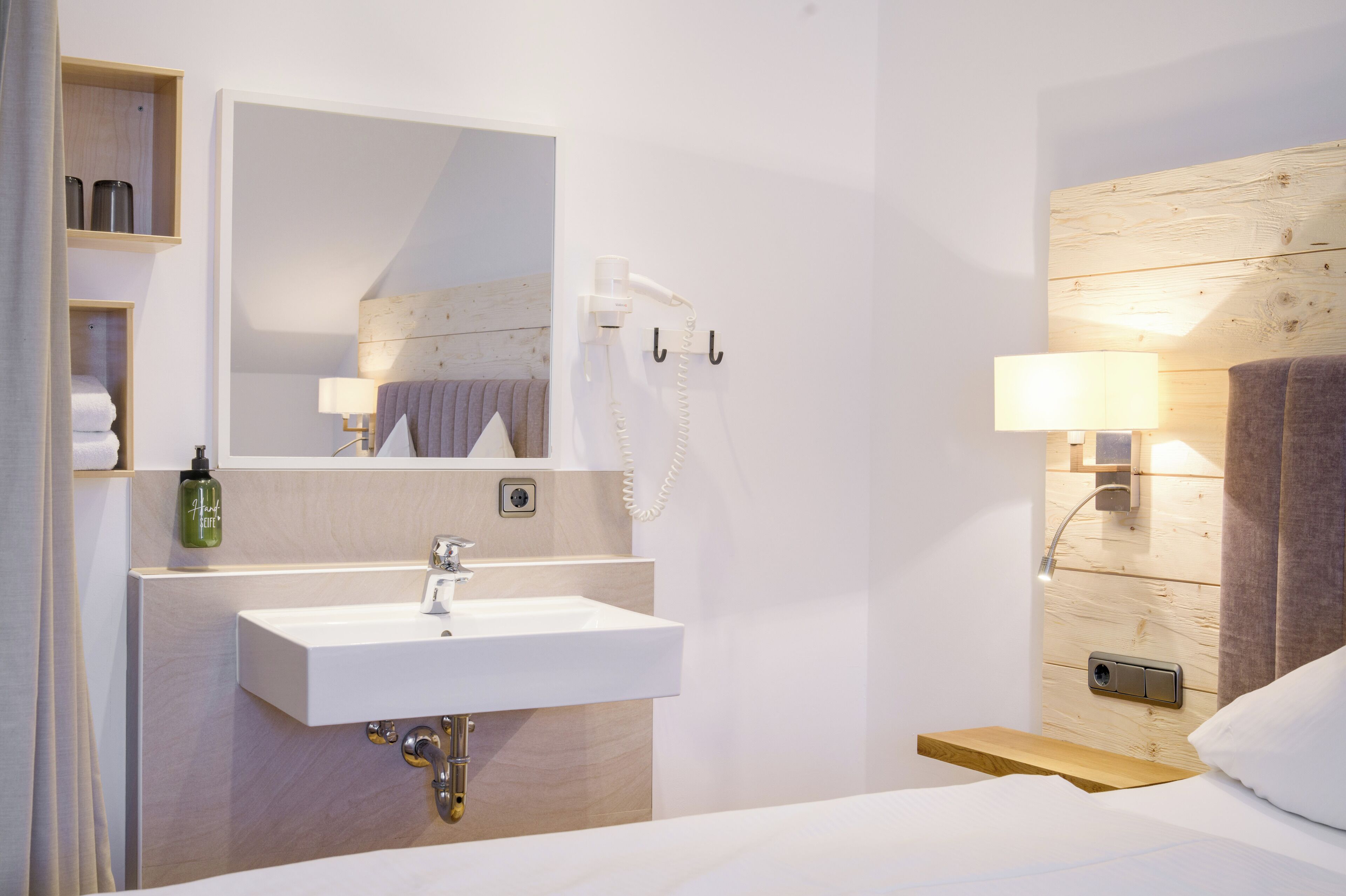A modern hotel room with a white sink, a mirror, a wooden headboard, a bedside table, and a lamp.