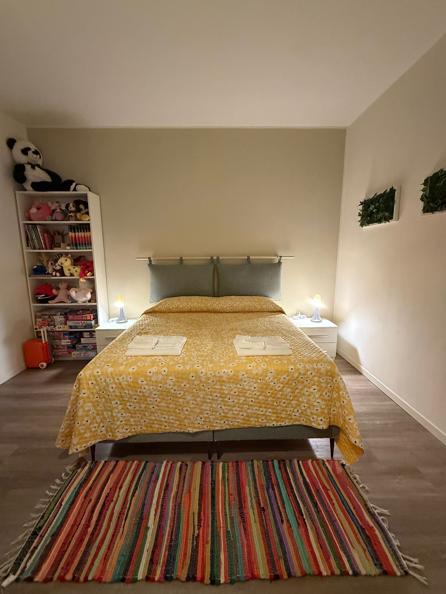 A bedroom with a bed, a colorful striped rug, a bookshelf with stuffed toys, and a nightstand with a lamp.