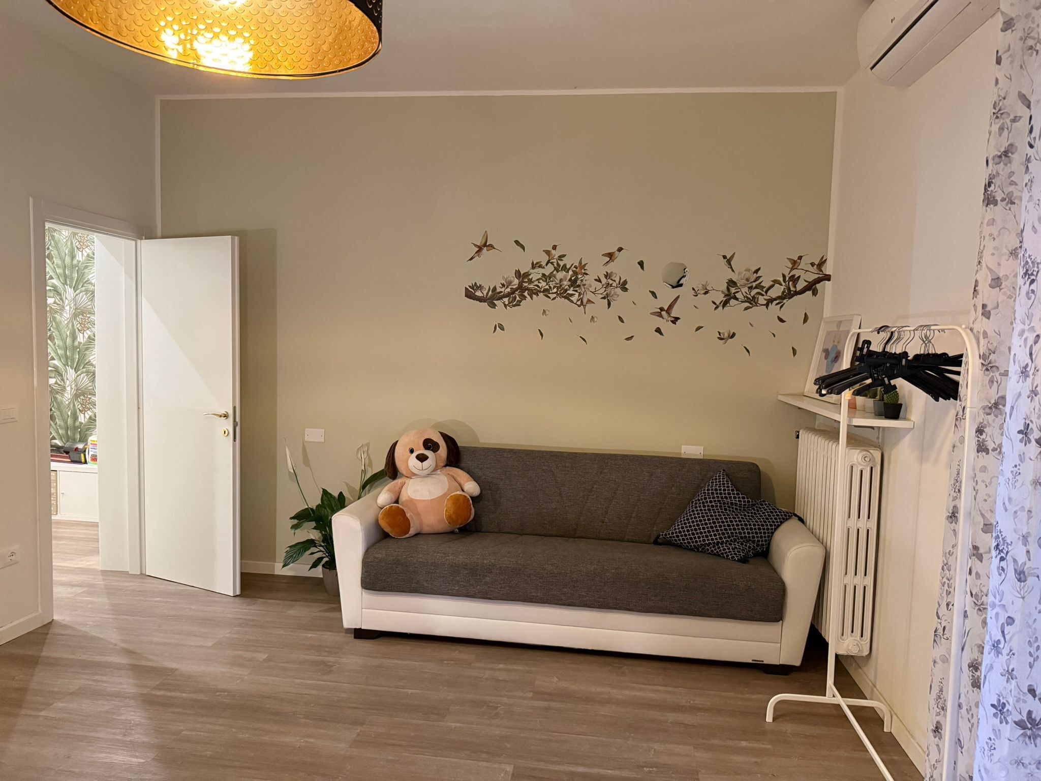 Un salon avec un canapé, un grand chien en peluche, un motif décoratif représentant des oiseaux sur un mur, un radiateur et une plante.
