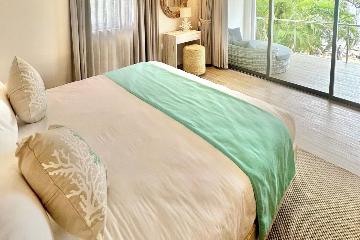 Une chambre spacieuse avec un grand lit, un ventilateur de plafond, un appareil de climatisation, une table de chevet avec une lampe, un miroir et une vue sur un balcon aménagé avec du mobilier d’extérieur.