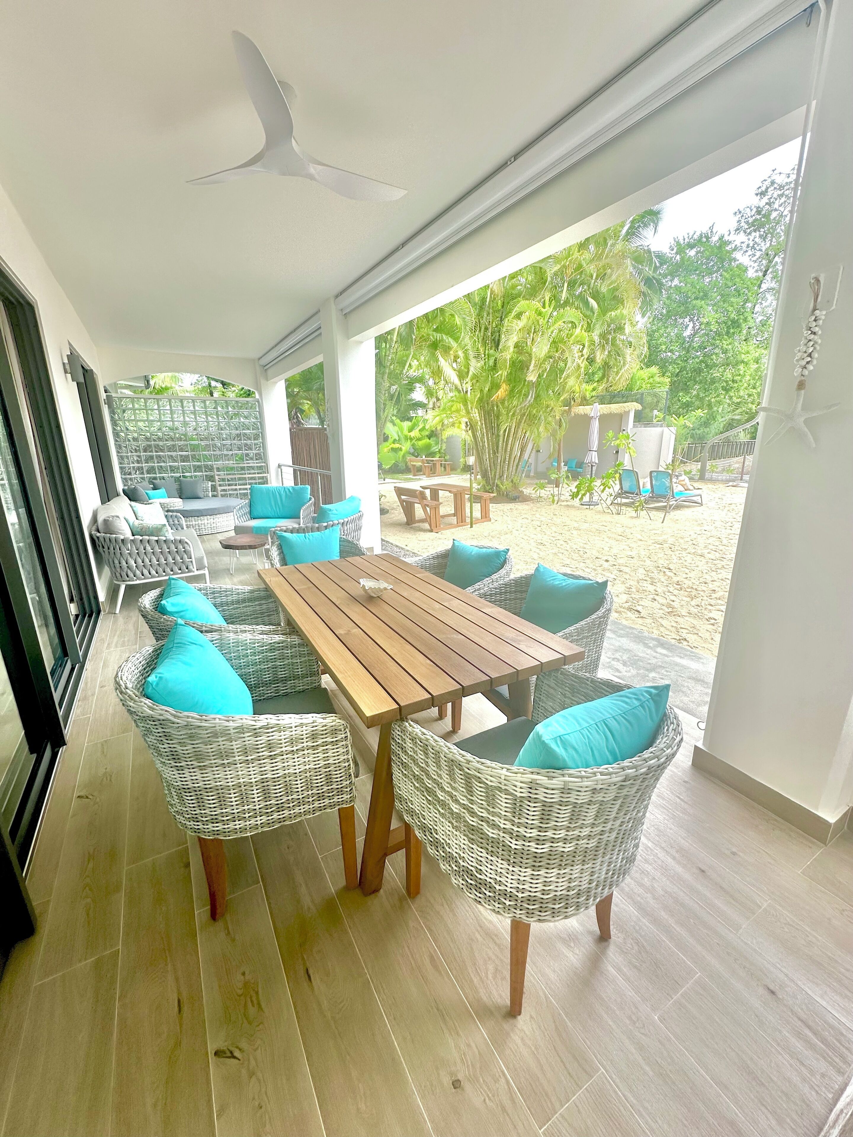 A covered outdoor seating area with wicker furniture and a wooden table, overlooking a sandy beach and lush greenery.