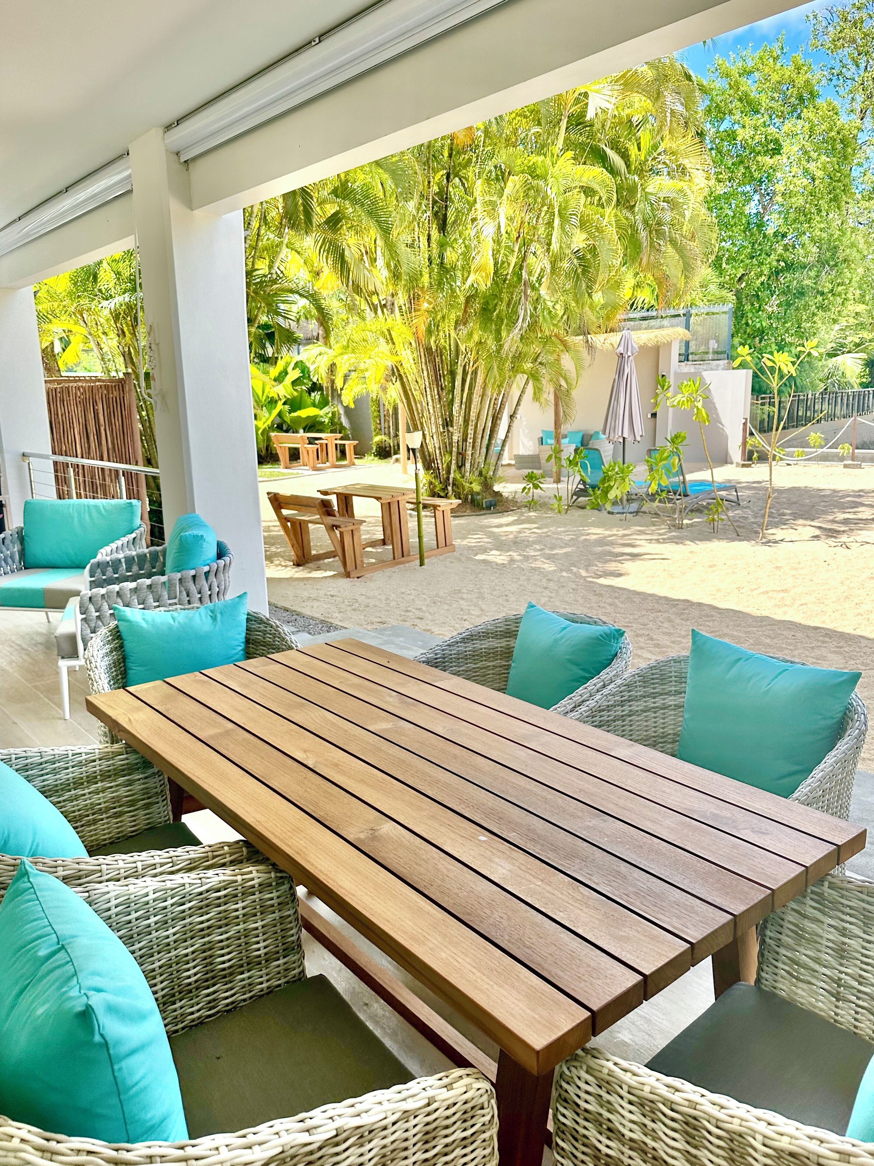 An outdoor patio area with a wooden table and wicker chairs, surrounded by greenery and a fence.