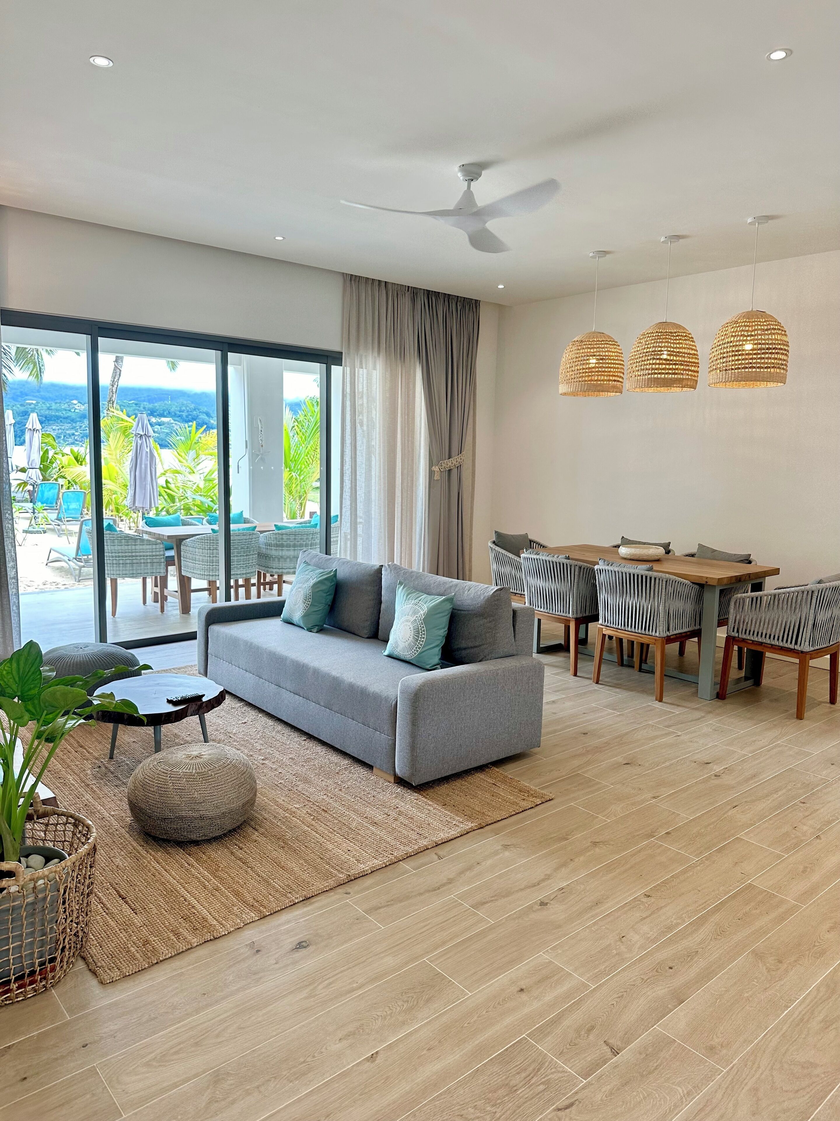 Una sala de estar moderna con un sofá gris, una mesa de comedor con sillas, un ventilador de techo y ventanas amplias que ofrecen vistas a zonas verdes y un paisaje lejano.