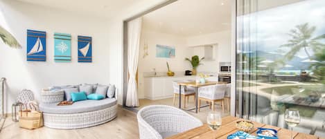 A modern living room with a curved sofa, a dining table with chairs, and a view of the kitchen and outdoor area through large windows.