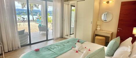Camera d'albergo con un letto grande, una ventola a soffitto, vista sulla spiaggia e una porta scorrevole in vetro.