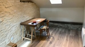 Una piccola stanza mansardata con soffitto spiovente, un tavolo di legno con una sedia, un velux e un muro in pietra.