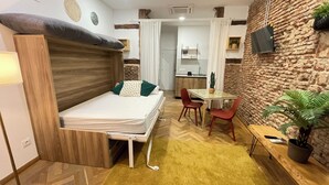 A compact room with a bed, a small dining table, red chairs, a potted plant, and a kitchen area. - WAKAFLATS 2 CHUECA SB (Madrid)