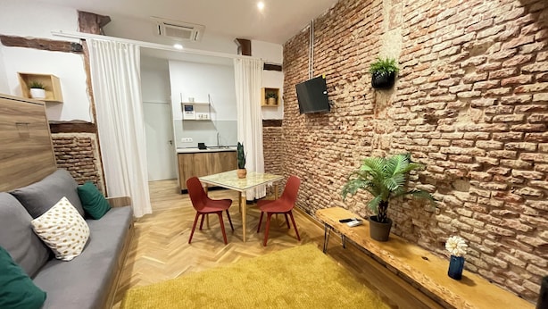A modern living room with a sofa, two red chairs, a glass table, and a yellow rug. A kitchen with a stone wall and a dining area with a table and chairs are visible in the background. - WAKAFLATS 2 CHUECA SB (Madrid)