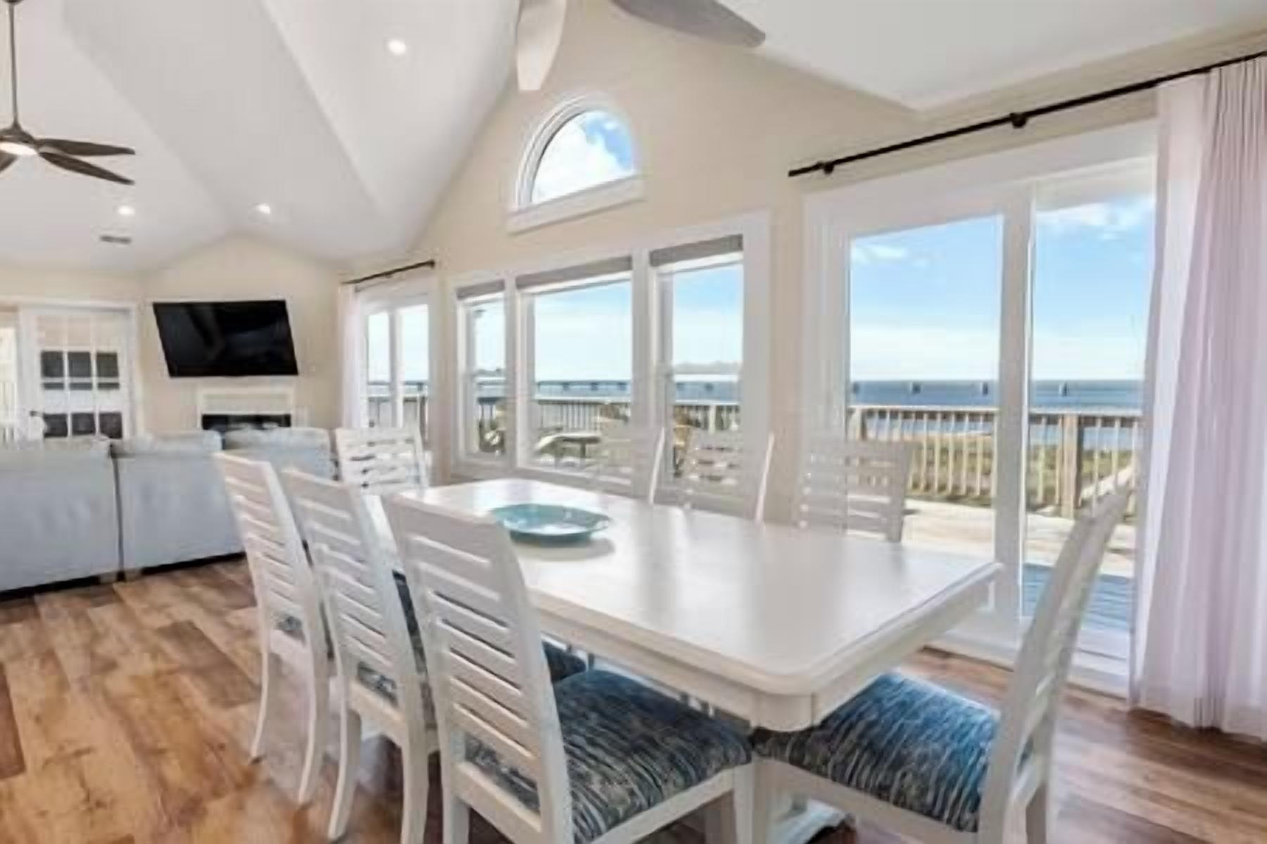 A spacious room with a large dining table, white chairs, and a view of the beach.