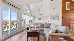 A spacious living room with a large window, a ceiling fan, and a wooden accent wall. - Luxury Sound-Front w/Ocean View (Rodanthe)