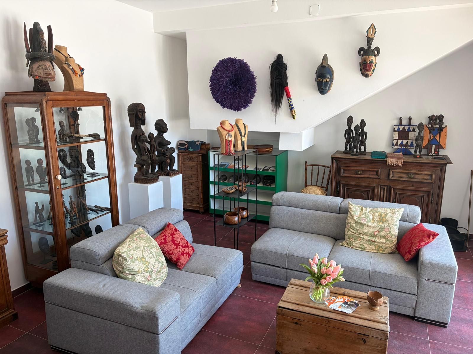 Una sala de estar con dos sofás grises, una mesa de centro de madera y una exhibición de artefactos culturales, incluyendo máscaras y esculturas.