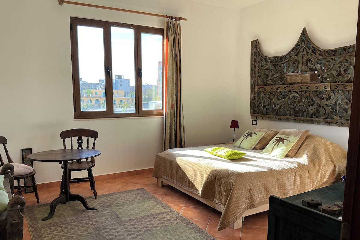 Quarto com cama, cadeira, mesa, janela e cabeceira decorativa.