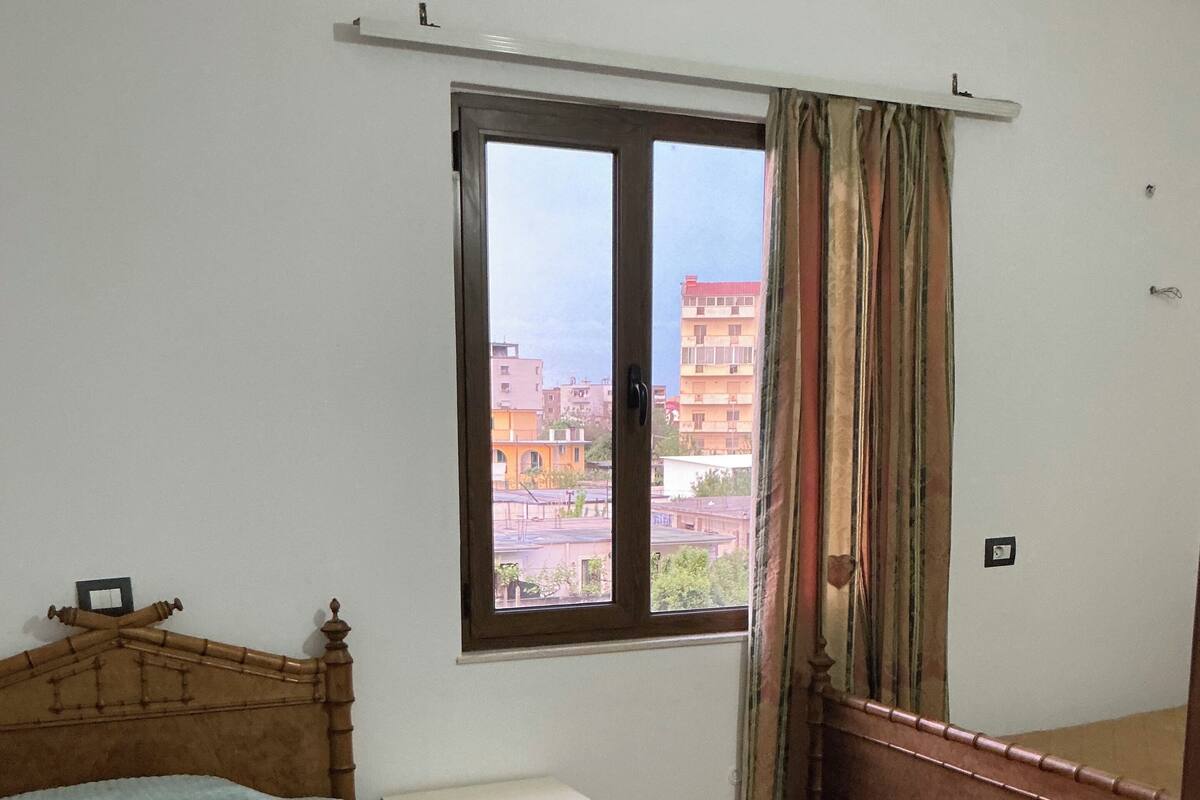 Quarto com cama, mesa de cabeceira, cômoda, janela com cortinas e vista para edifícios.