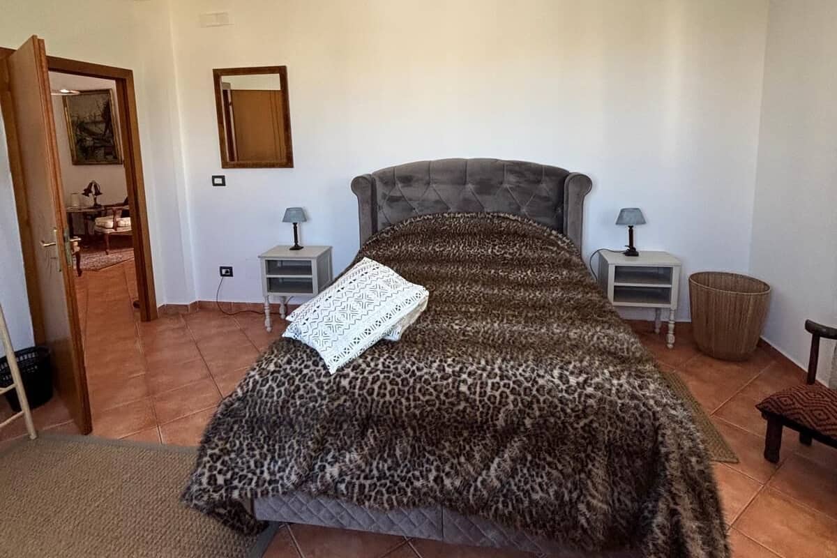 Quarto com cabeceira estofada, colcha estampada, duas mesas de cabeceira com luminárias, uma porta que dá para outro cômodo e um lustre de palha no teto.