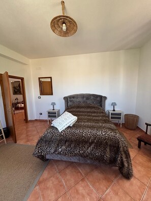 A bedroom with a tufted headboard, a patterned bedspread, two bedside tables with lamps, a door leading to another room, and a woven ceiling light fixture. - Hostel Calao (Shkoder)