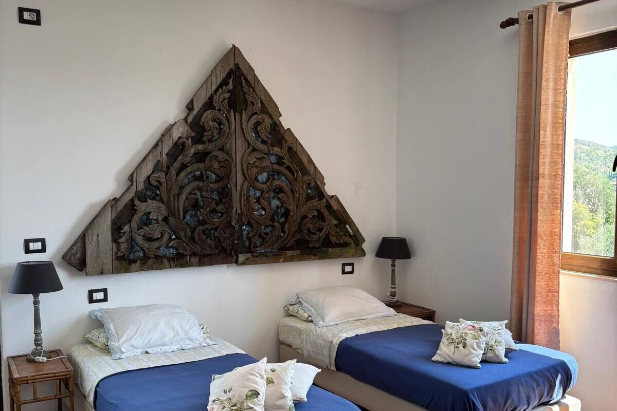 Quarto com duas camas, uma cabeceira de madeira decorativa, um candeeiro vermelho e uma janela com vista para o verde.