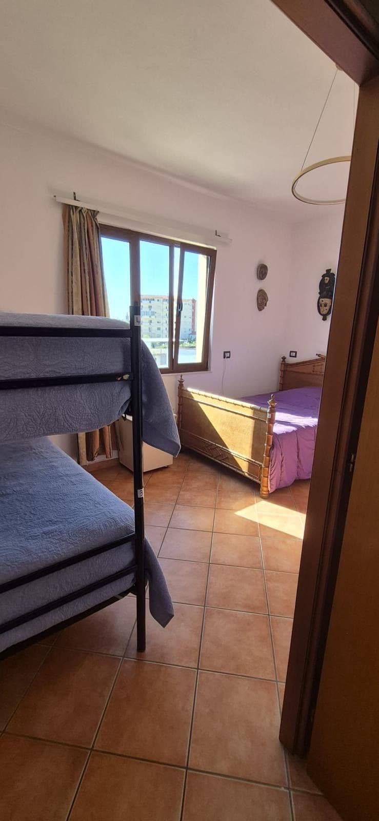 A room with a bunk bed, a single bed, a window with curtains, and a view of buildings outside.