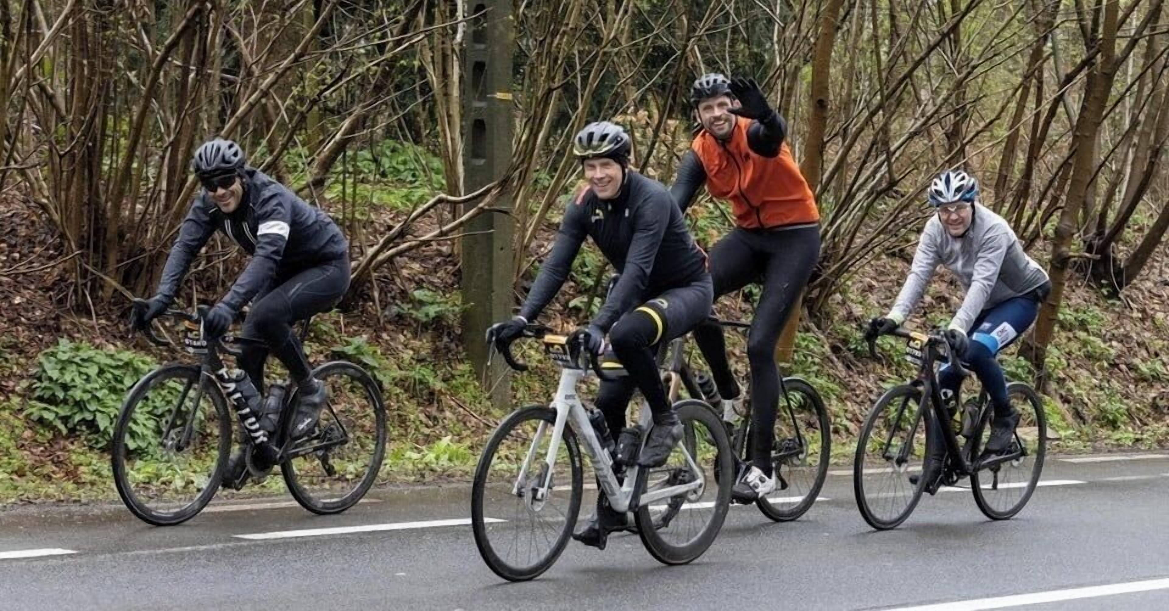 Four cyclists on a road with trees and a fence in the background.