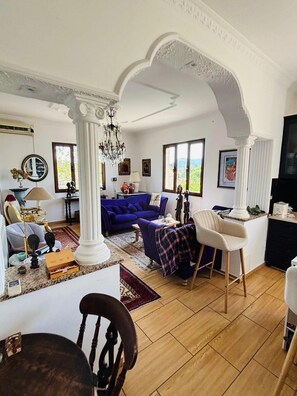 Ein geräumiges Wohnzimmer mit einem blauen Sofa, einer Kristallleuchter und Blick durch große Fenster.