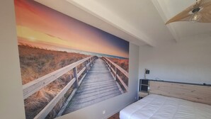 Un dormitorio con piso de madera, una cama blanca, un escritorio de madera y un mural en la pared que muestra un muelle de madera que lleva a una playa al atardecer.