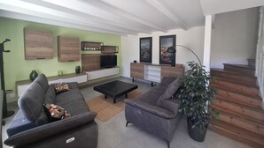 Una sala de estar moderna con un sofá gris, una mesa de centro y un televisor sobre un mueble de madera. A la derecha se encuentra una escalera y una pared verde con obras de arte.