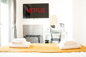 Habitación de hotel con una cama, un televisor en la pared mostrando "NETFLIX", un escritorio con espejo y una cafetera.