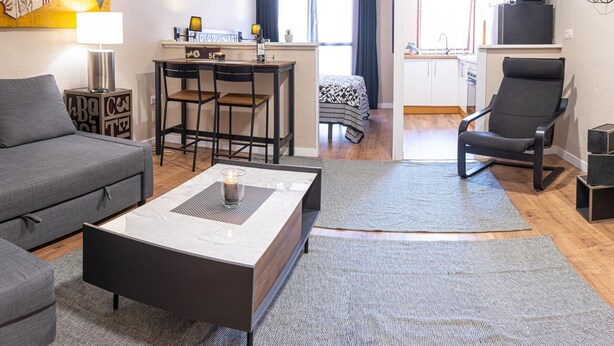 A modern living room with a grey sofa, a coffee table, and a dining area with a table and chairs. There is a kitchen visible in the background with a refrigerator and a microwave. - Elegant Loft in Torremolinos Center with Pools, Near Beach and Nightlife (Torremolinos)