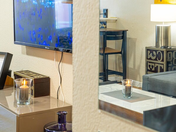 Habitación de hotel con televisión de pantalla plana en la pared, una mesita con una vela, un sofá y una caja decorativa con la palabra 'HOME'.