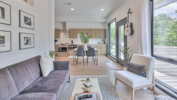 A modern living room with a grey sofa, a coffee table, and a dining area with a table and chairs. There is a kitchen in the background with wooden cabinets and a large window.