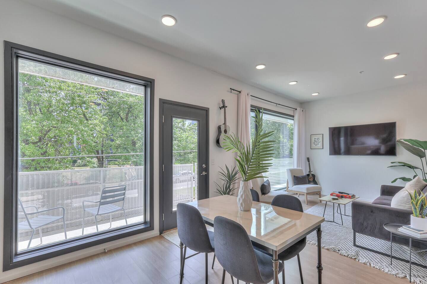 Un salon moderne comprenant un coin repas, de grandes fenêtres, un téléviseur à écran plat et un balcon aménagé avec du mobilier d’extérieur.