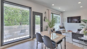 A modern living room with a dining area, large windows, a flat-screen TV, and a balcony with outdoor furniture.