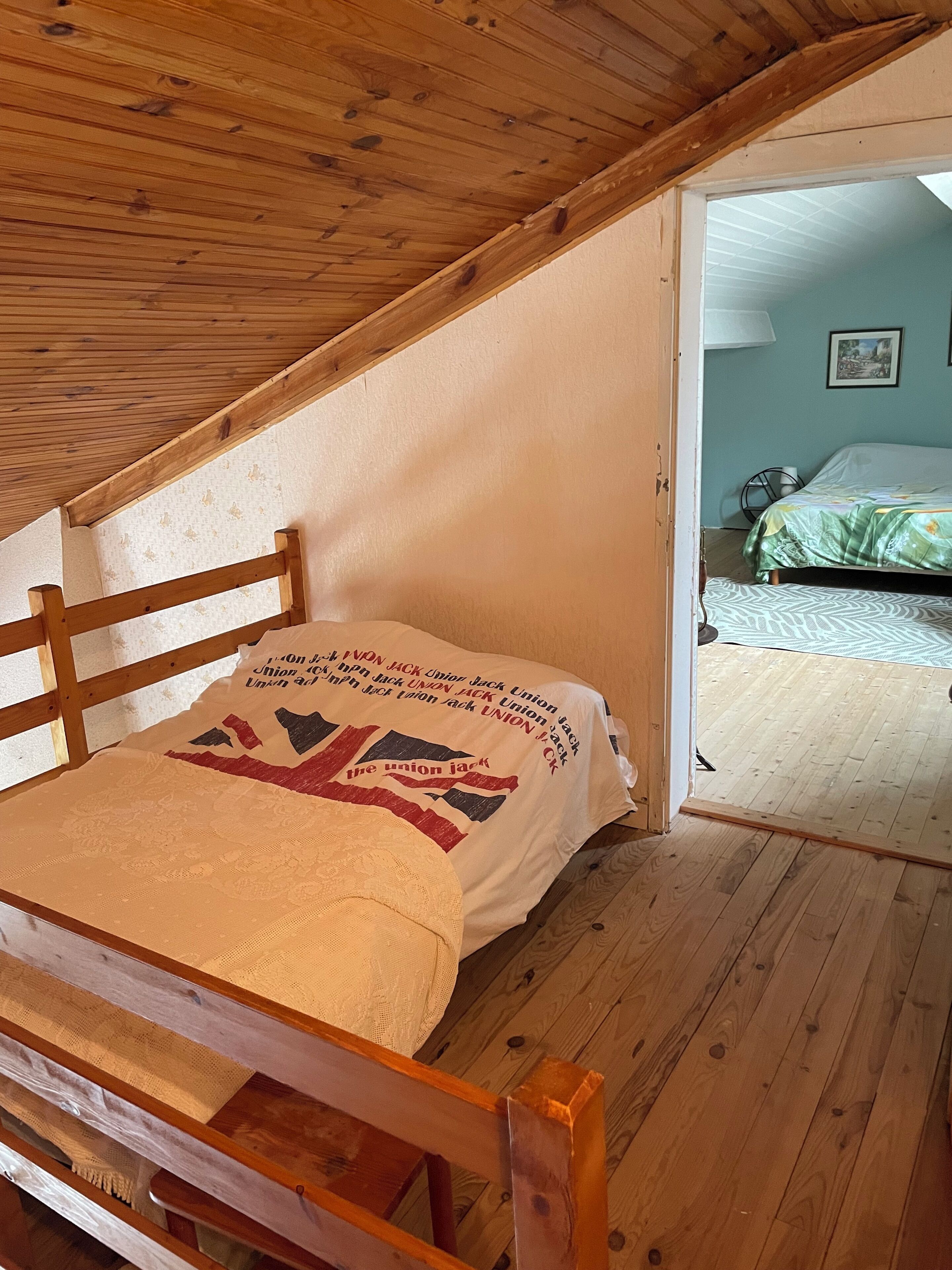 Une chambre avec un lit superposé en bois, un lit simple orné d’un coussin aux couleurs de l’Union Jack, et une porte donnant sur une autre pièce.