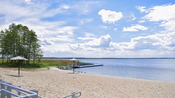 A beach with a wooden pier extending into calm waters, surrounded by sand and sparse vegetation.