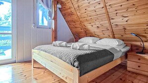A wooden bed in a room with a sloped ceiling, a window with curtains, and a nightstand with a lamp.