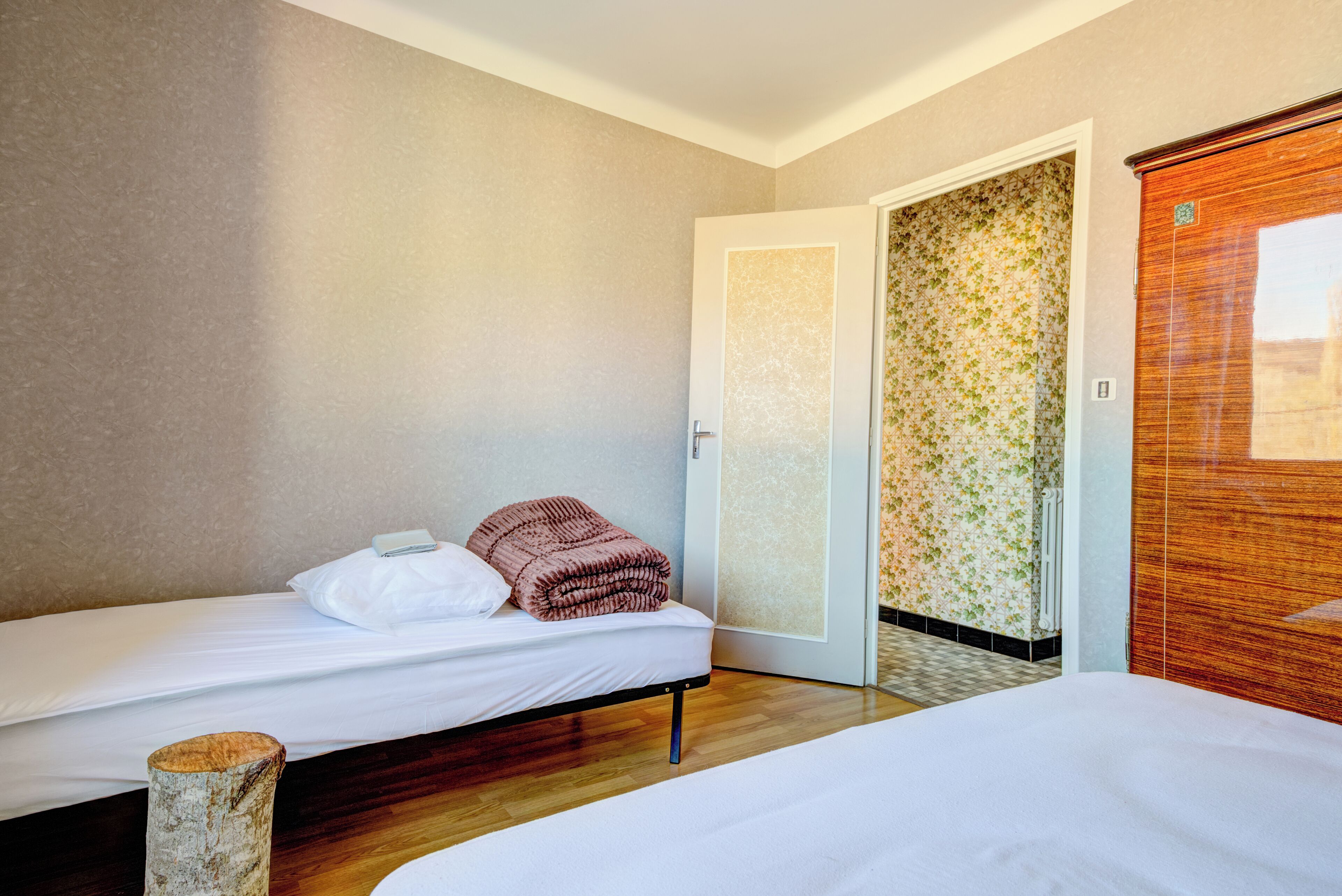 Une chambre d’hôtel avec deux lits simples, un plancher en bois, une porte recouverte d’un papier peint à motifs floraux et une armoire en bois.